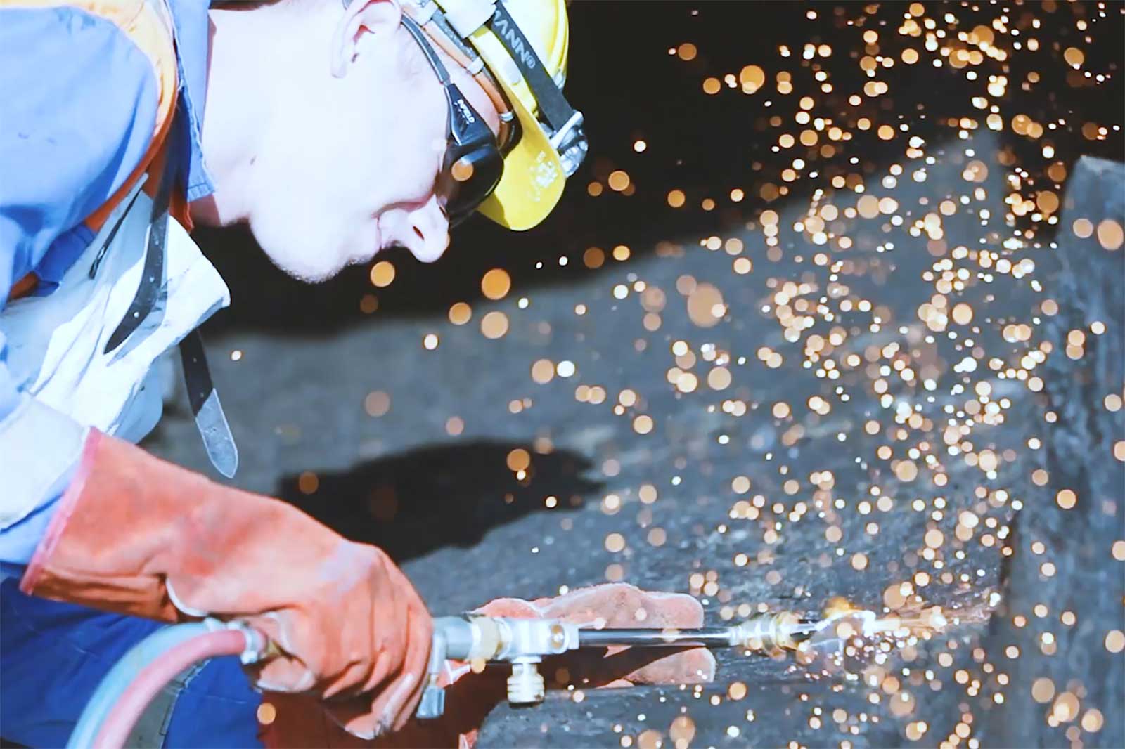 Ein Arbeiter in Schutzausrüstung führt Schweißarbeiten durch und erzeugt helle Funken vor einem dunklen Hintergrund.