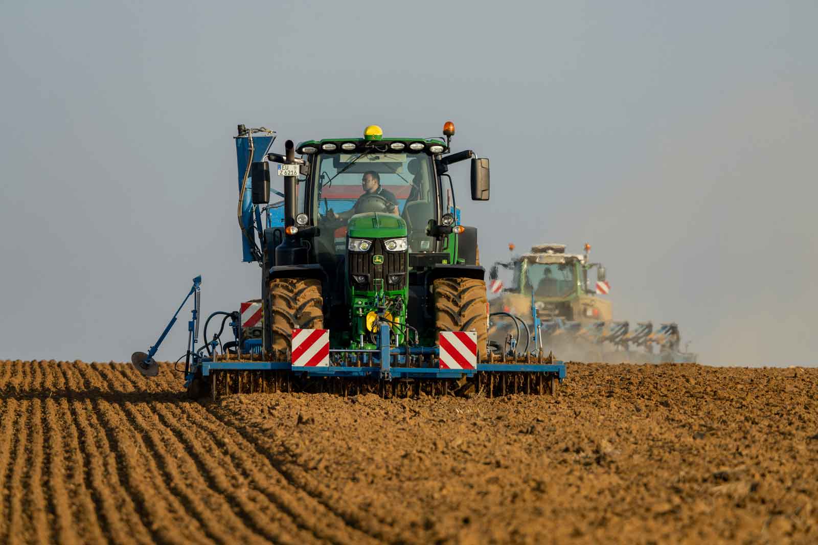Ein grüner Traktor pflügt ein Feld mit frischer Erde, ein weiterer Traktor im Hintergrund sichtbar.