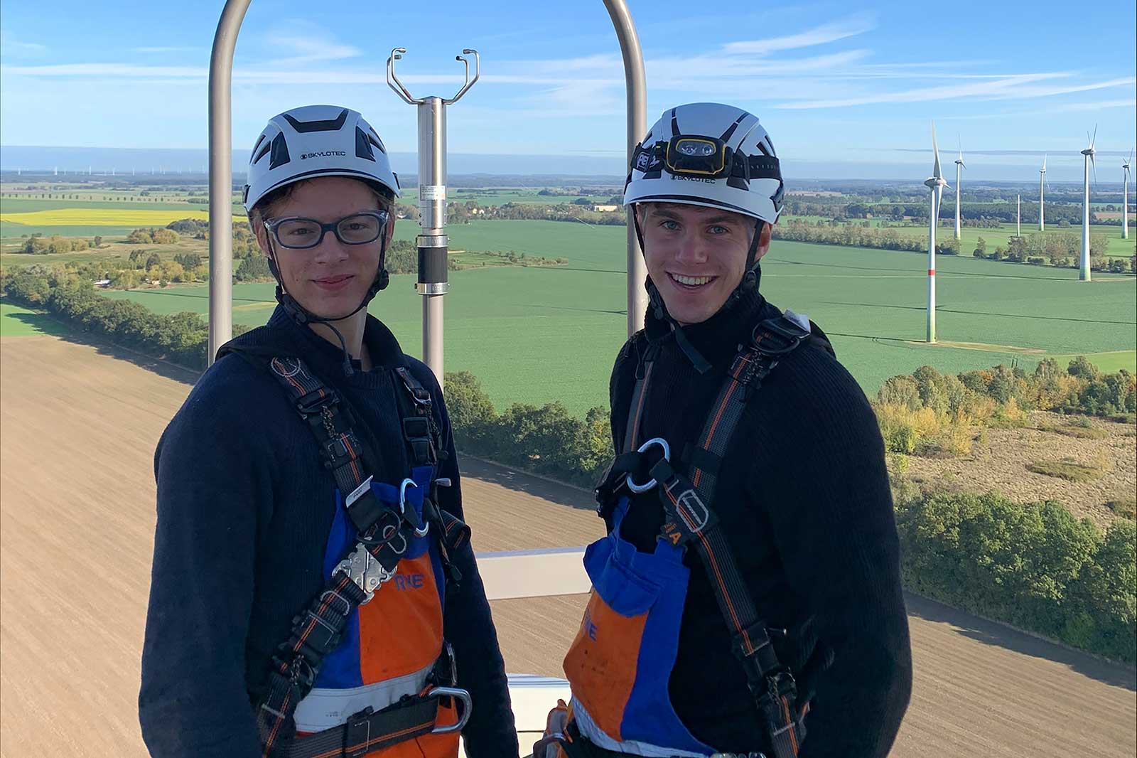 Zwei Personen in Sicherheitsausrüstung stehen auf einer Plattform über einer grünen Landschaft mit Windturbinen im Hintergrund.