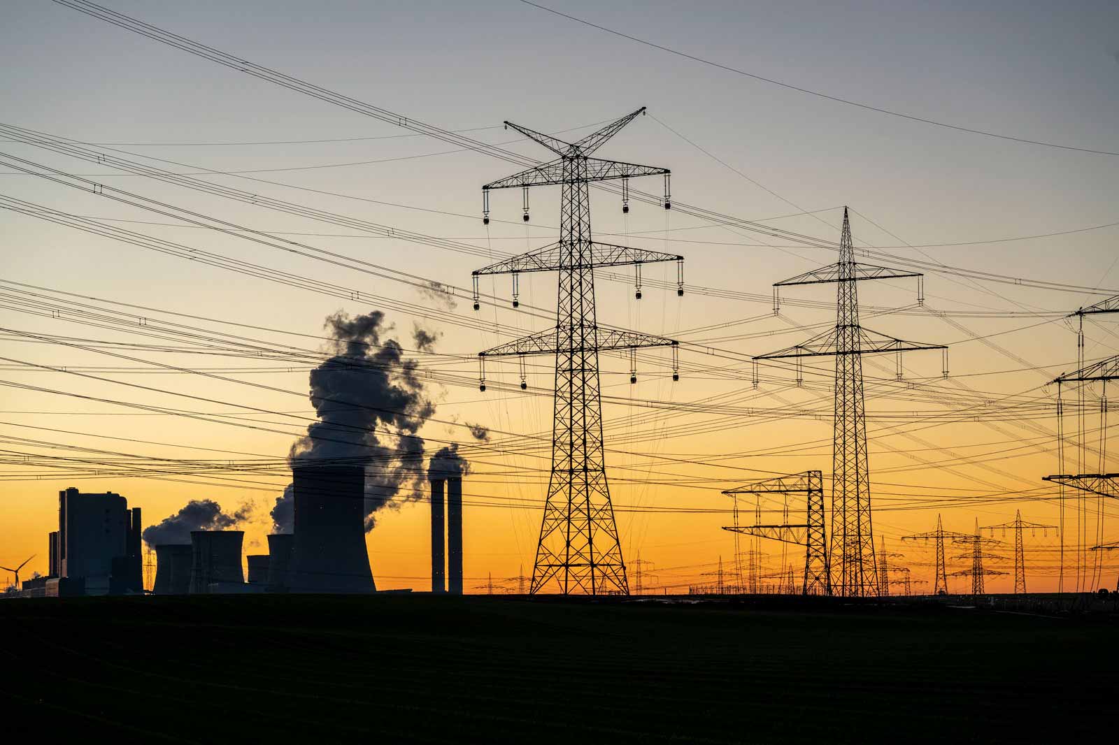 Silhouettierte Stromleitungen vor einem beeindruckenden Sonnenuntergang mit Rauch, der von Industrieanlagen aufsteigt.