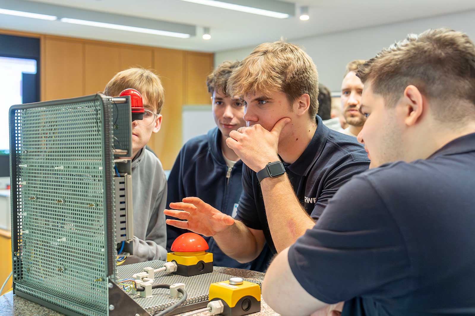 Eine Gruppe von Personen diskutiert über Steuerungskomponenten, einschließlich eines großen roten Knopfs und verschiedener Schalter.