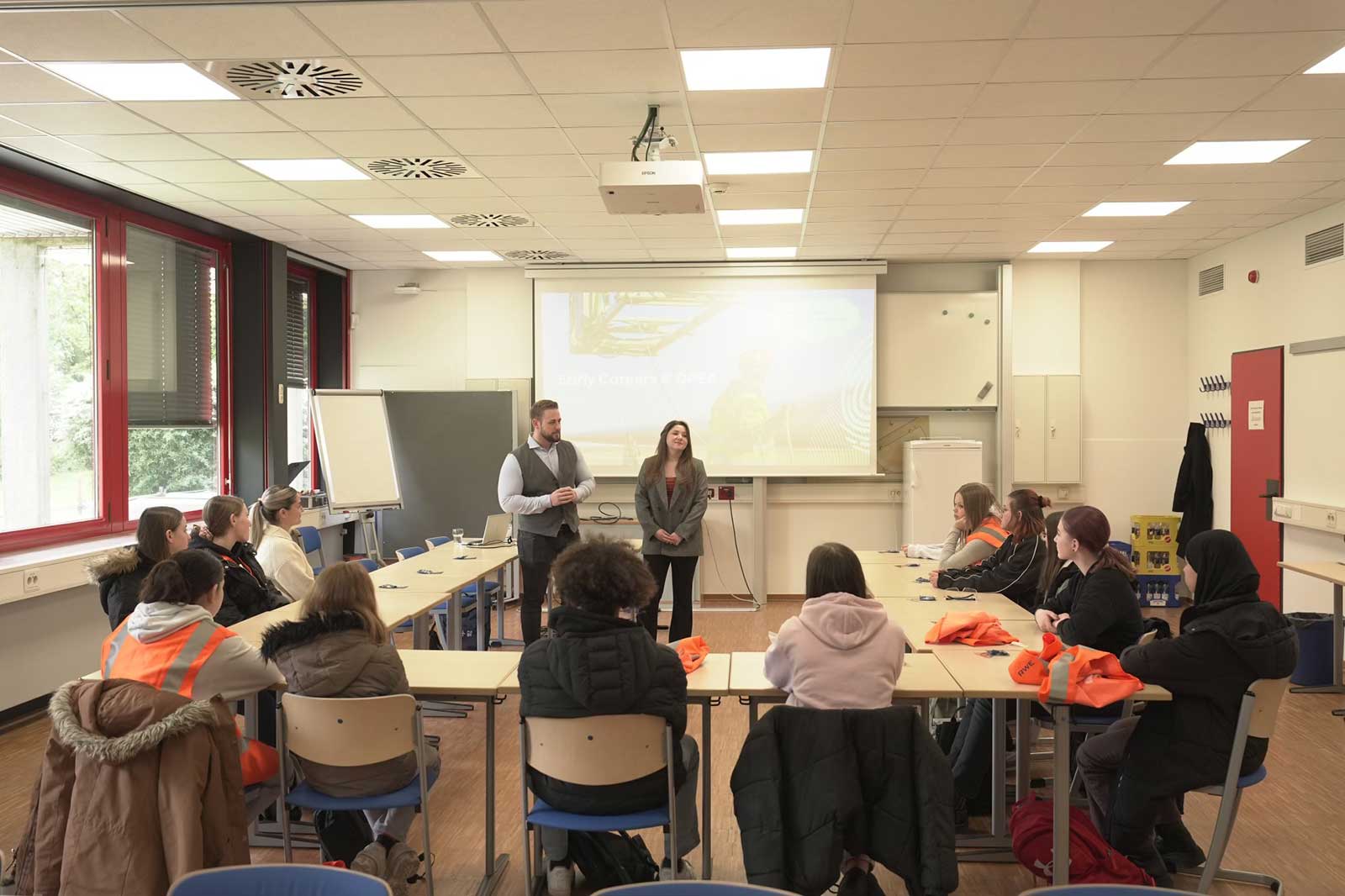 Eine Klassenzimmersituation mit Teilnehmenden, die an einem Tisch sitzen. Zwei Referenten präsentieren vor einem Bildschirm mit Bildern.