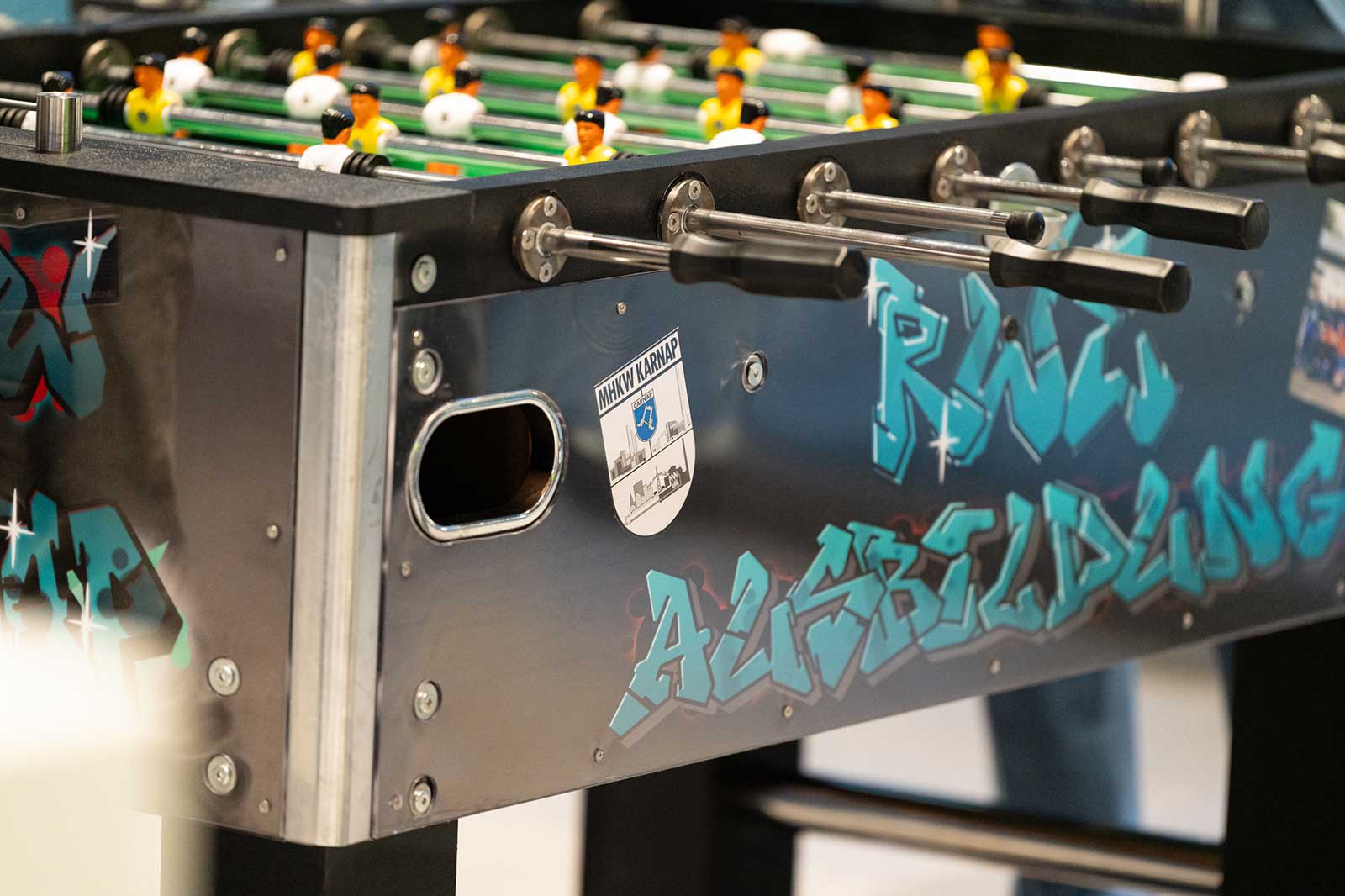 Ein Tischfußballspiel mit bunten Spielerfiguren und Graffiti-ähnlichem Text auf der Oberfläche, in einer lebhaften Umgebung.