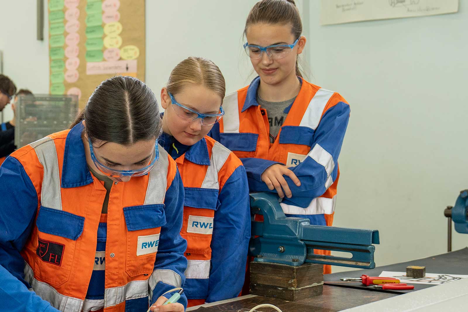 Eine Gruppe von Auszubildenden in Sicherheitskleidung arbeitet gemeinsam an einem Werkstatttisch mit Werkzeugen und Geräten.