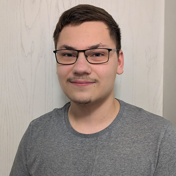 Eine Person in einem grauen T-Shirt, die vor einer hellen Holzwand steht, mit einem neutralen Ausdruck.