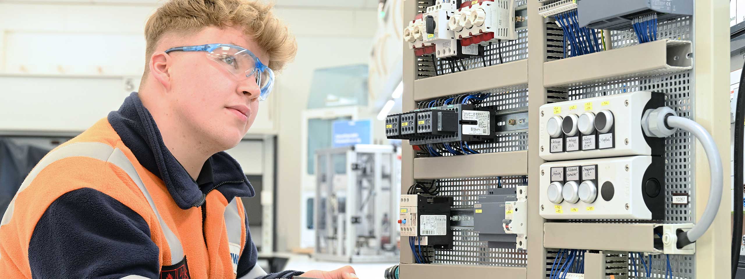Ein Techniker in orangefarbener Warnkleidung arbeitet an einem Schaltkasten mit verschiedenen elektrischen Komponenten.