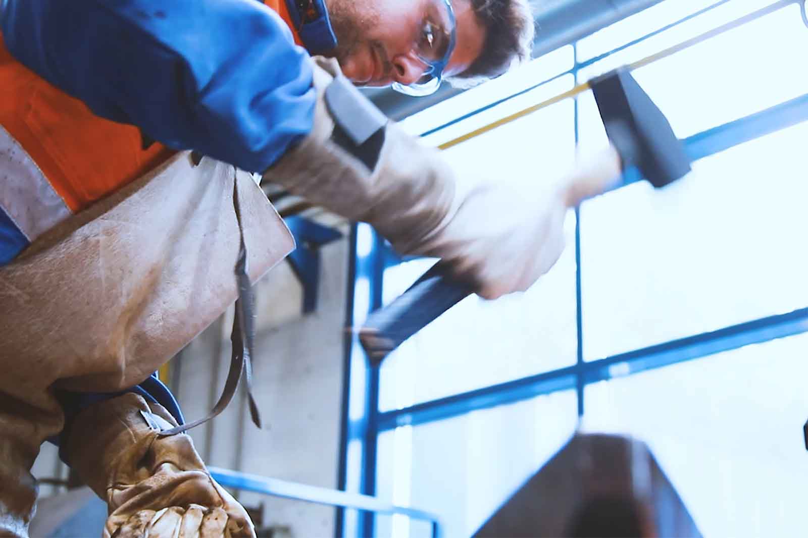 Eine Person in Arbeitshandschuhen schlägt mit einem Hammer auf einen Gegenstand und trägt eine blaue und orange Arbeitjacke in einer Werkstatt.