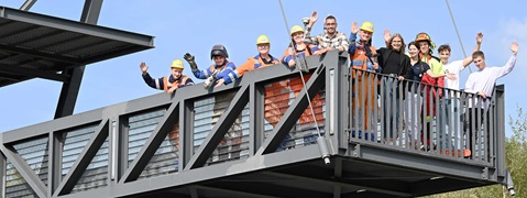 Eine Gruppe von Arbeitern in Sicherheitskleidung, die von einer Metallplattform vor einem klaren blauen Himmel winken.