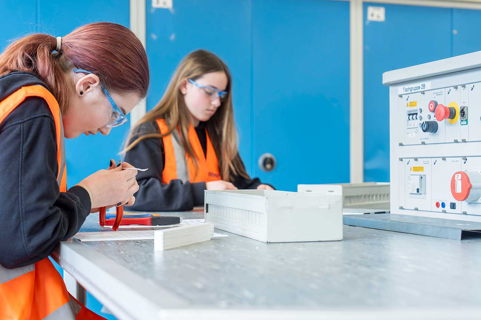 Zwei Personen arbeiten an einem Tisch mit Werkzeug und Materialien in einer technischen Umgebung. Die Wände sind blau.