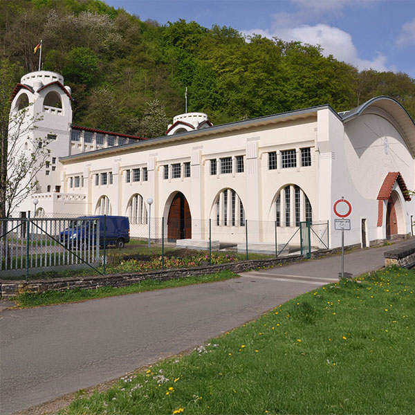 Ein cremefarbenes Gebäude mit Rundtürmen und großen Bogenfenstern steht hinter einem Metallzaun an einer Straße vor bewaldetem Hügel unter blauem Himmel.
