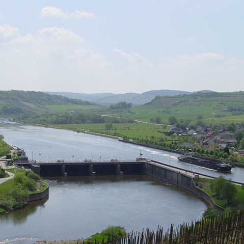 Eine breite Beton-Schleusenanlage quert den ruhigen Fluss, rechts liegt ein dunkler Frachter, im Hintergrund grüne Hügel, Wiesen und ein kleines Dorf.