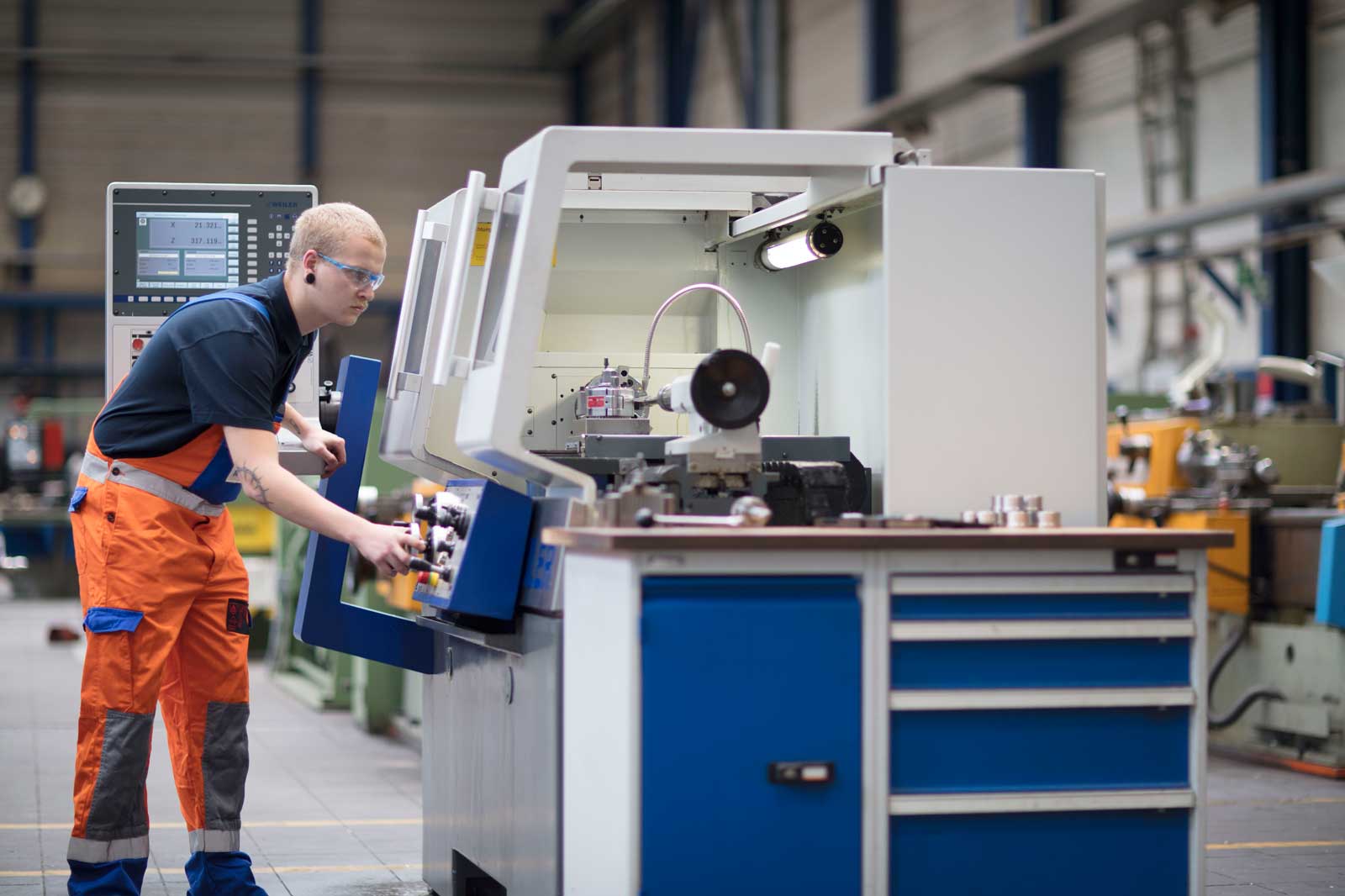 Eine Person, die in einer Werkstatt eine Maschine bedient, trägt orangefarbene Overalls und arbeitet mit mechanischen Komponenten.