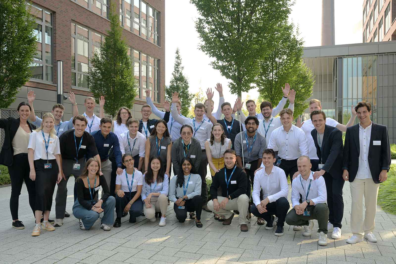 A diverse group of people posing together outdoors, smiling and waving, surrounded by trees and modern buildings.
