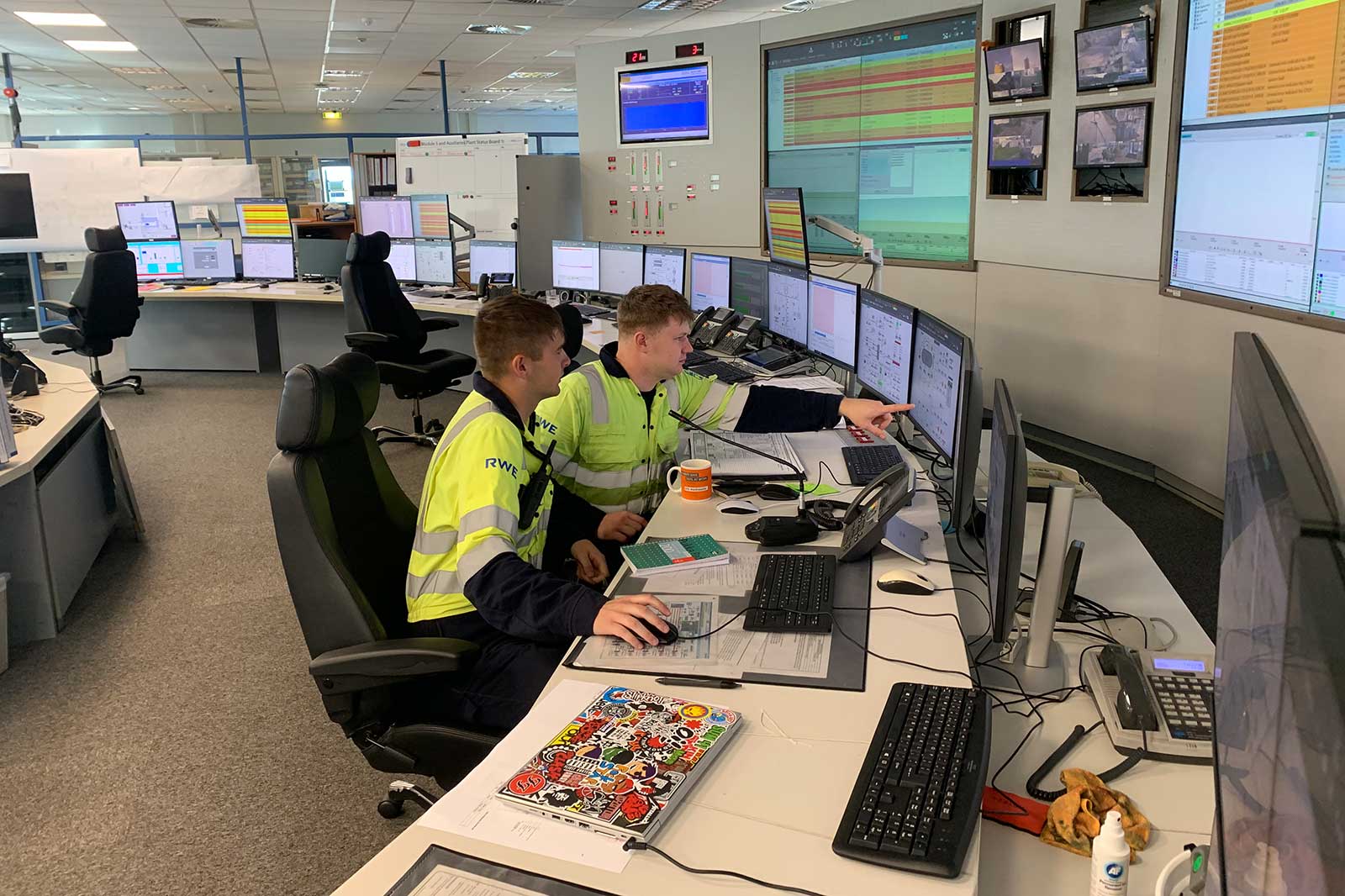 Two men in work uniforms focus on multiple computer screens in a control room, with documents and a coffee mug on the desk.