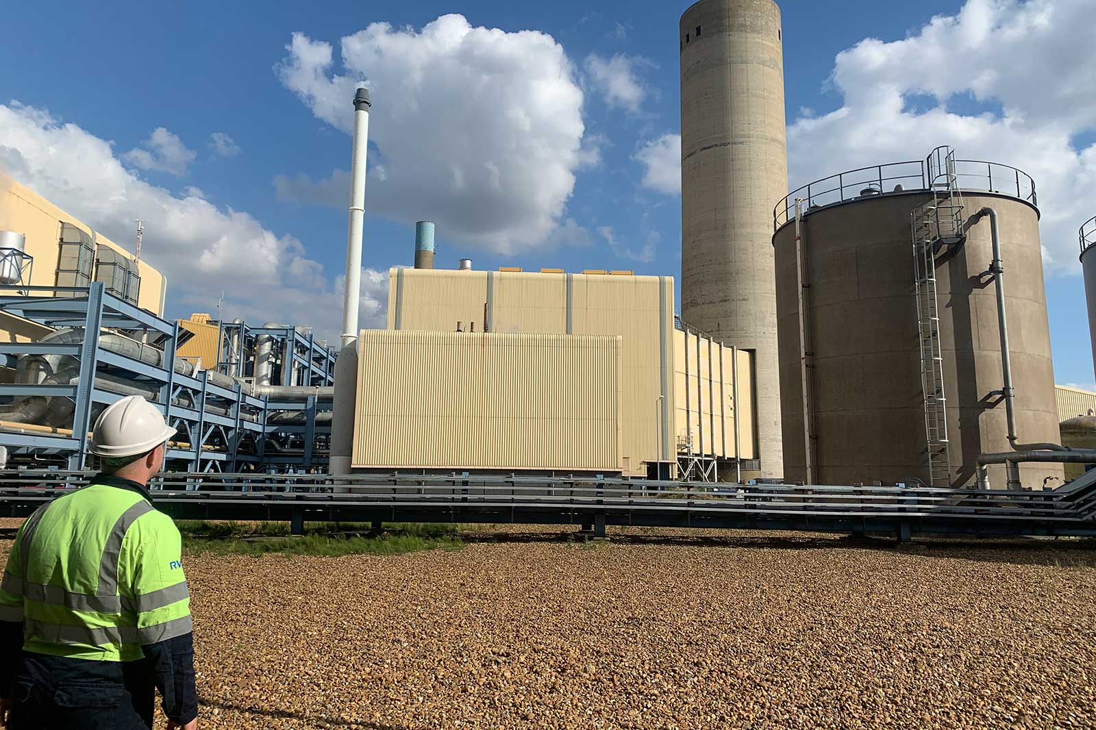 A worker in a hard hat stands before an industrial facility with several large silos and pipes under a blue sky.