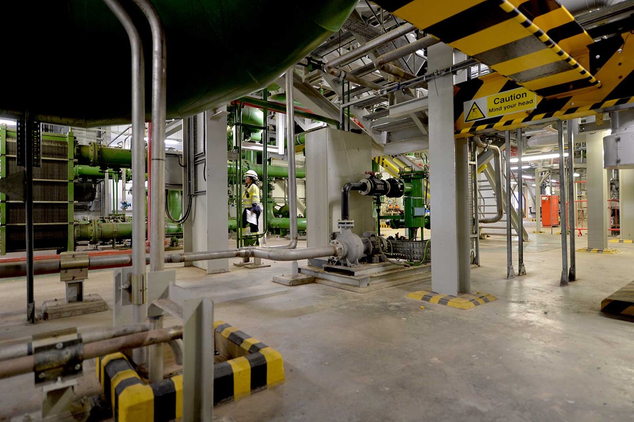 An industrial facility featuring green pipes, a large pump, and a person in a hard hat walking through the space.