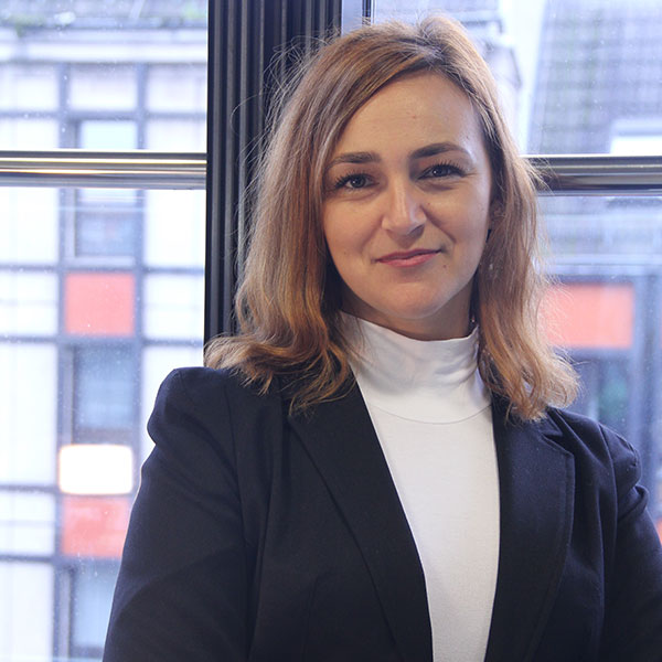 Eine Frau mit langen braunen Haaren trägt ein schwarzes Sakko und einen weißen Rollkragenpullover und steht an einem Fenster mit verschwommenem Stadtblick.