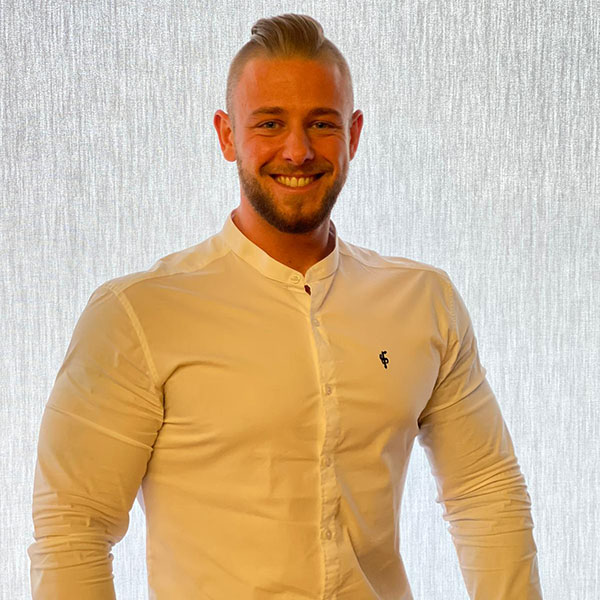 Ein Mann trägt ein weißes Hemd mit einem kleinen Logo. Er steht vor einem hellen, strukturierten Hintergrund.