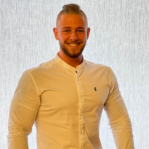A man is wearing a white shirt with a small logo. He is standing in front of a light, textured background.