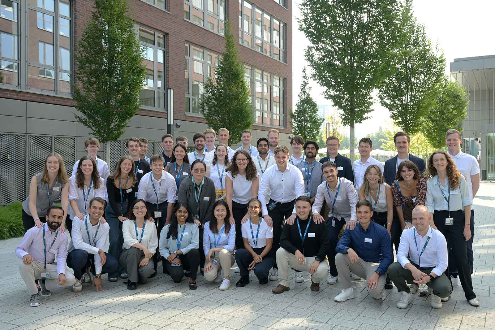 A group of diverse individuals gathered outdoors, standing in front of a modern brick building with trees.