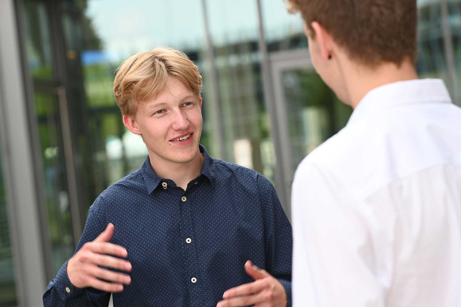Zwei junge Männer führen im Freien ein Gespräch. Ein Mann trägt ein dunkelblaues Hemd mit weißen Punkten, der andere ein weißes Hemd.