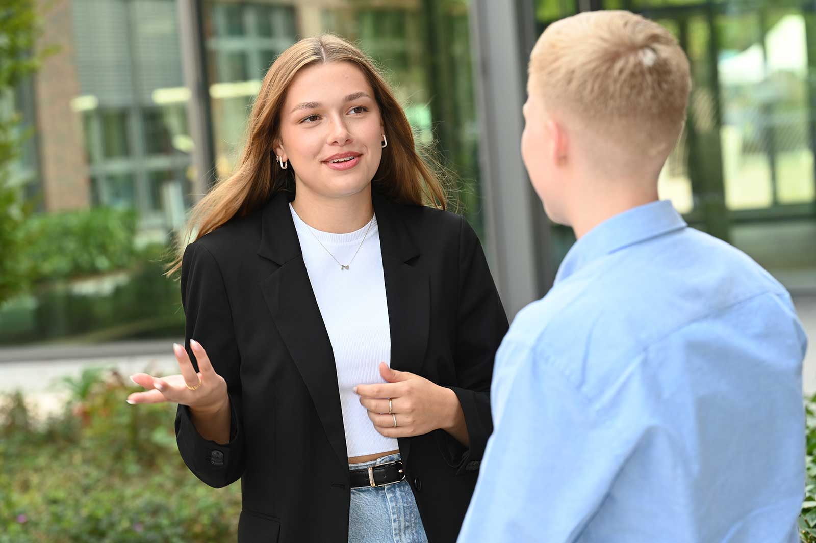 Eine Frau in einem schwarzen Blazer und einem weißen Oberteil kommuniziert lebhaft mit einem Mann in einem blauen Hemd im Freien.
