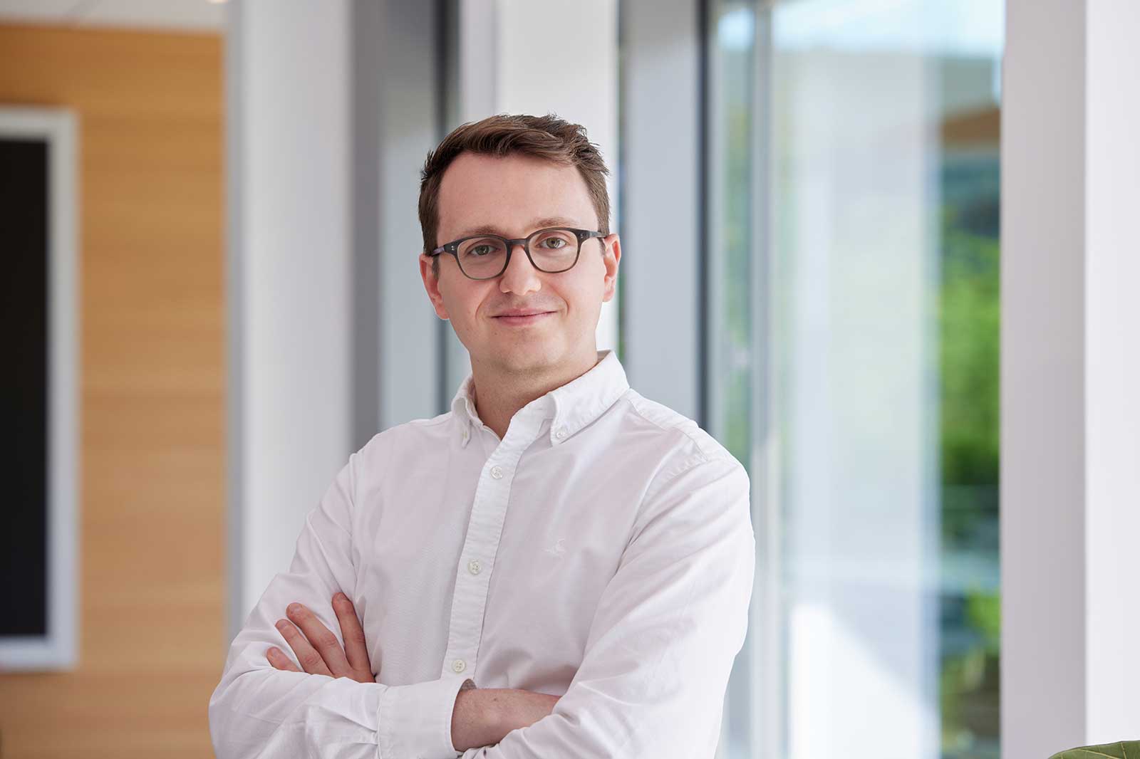 A person in a white shirt stands in a bright, modern office with glass windows and wooden accents.