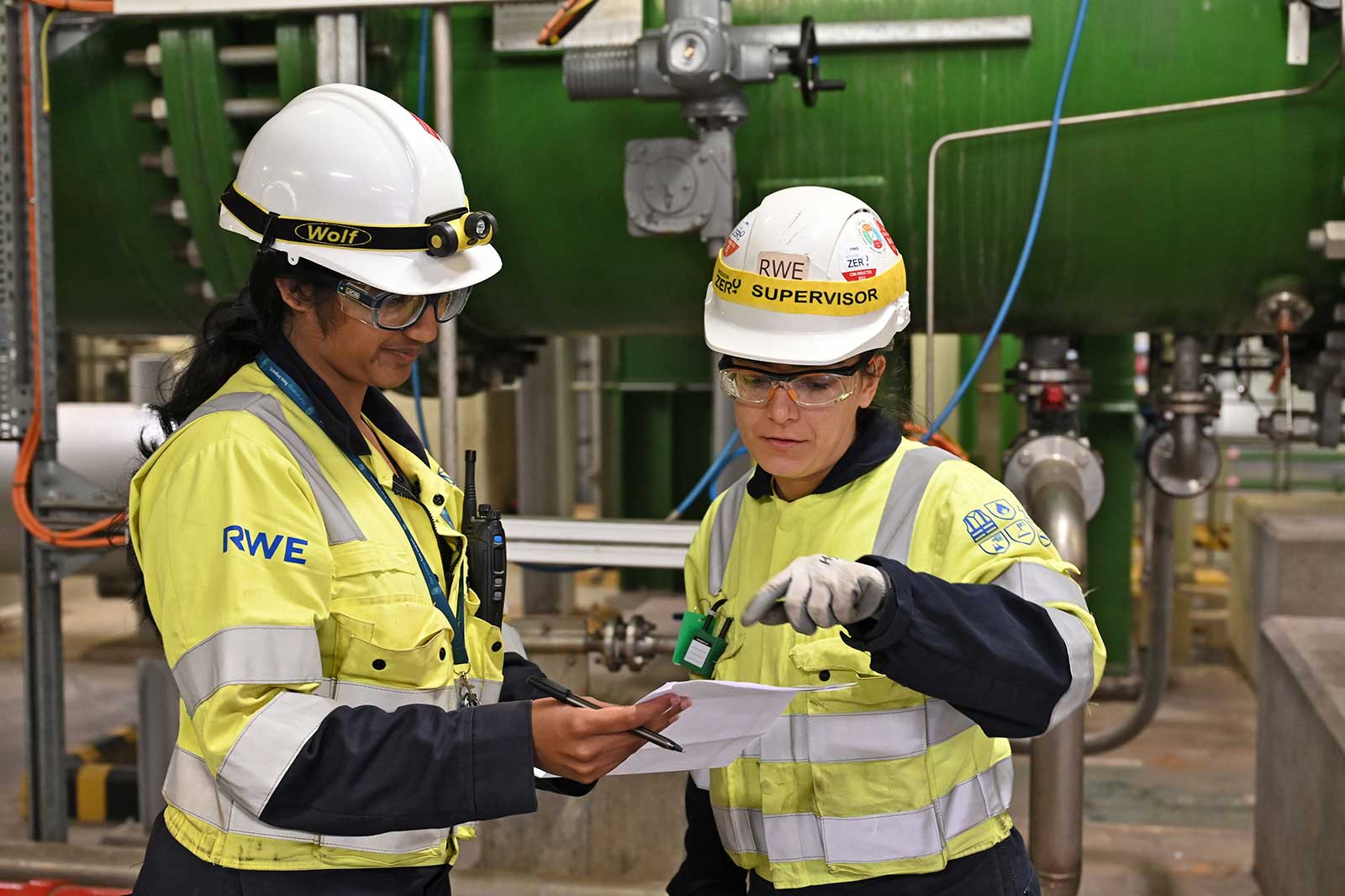 Two professionals in safety gear discuss documents near industrial equipment in a facility.