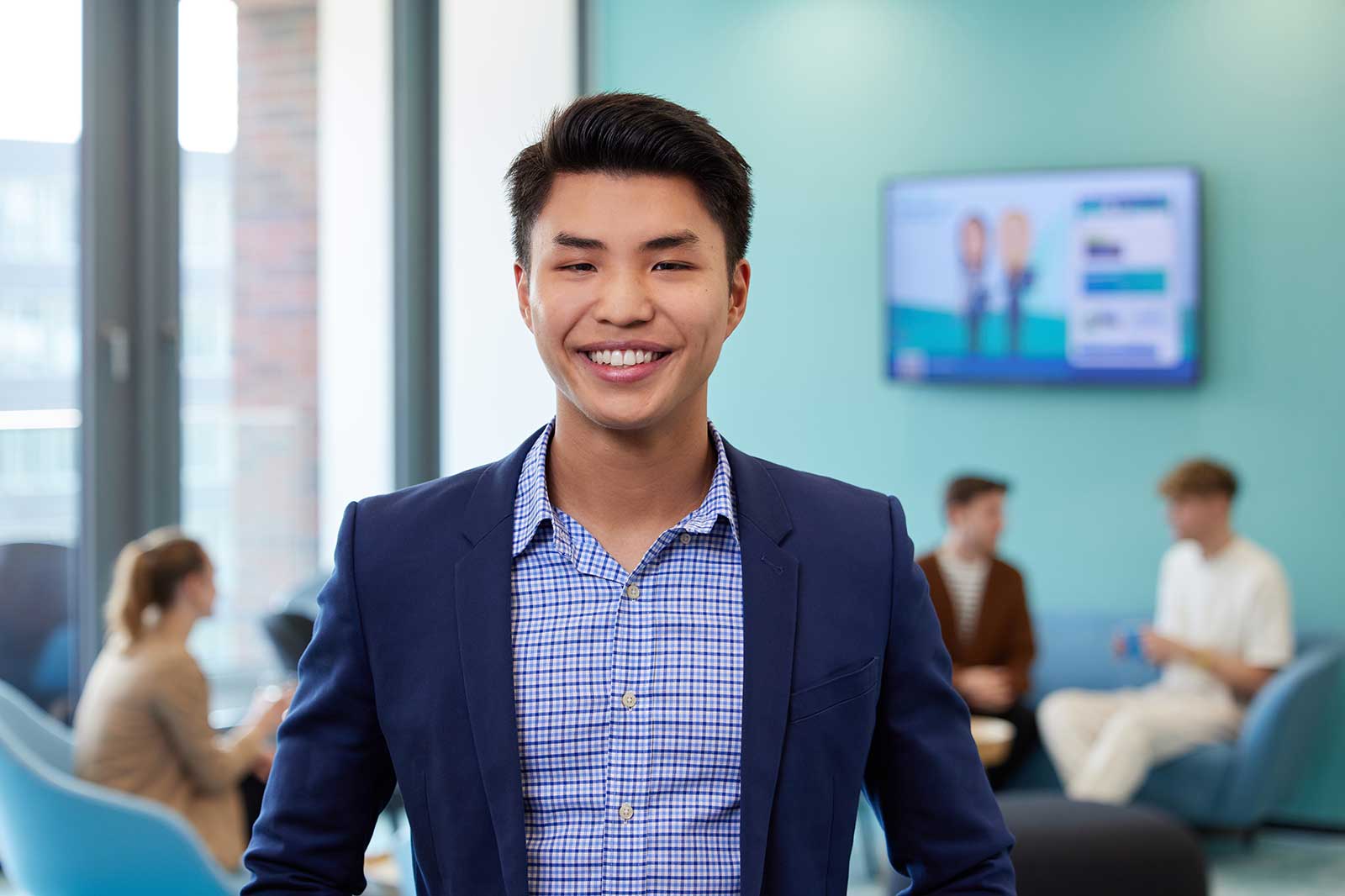 A businessman in a blue blazer stands in an office space, with blurred figures seated in the background.