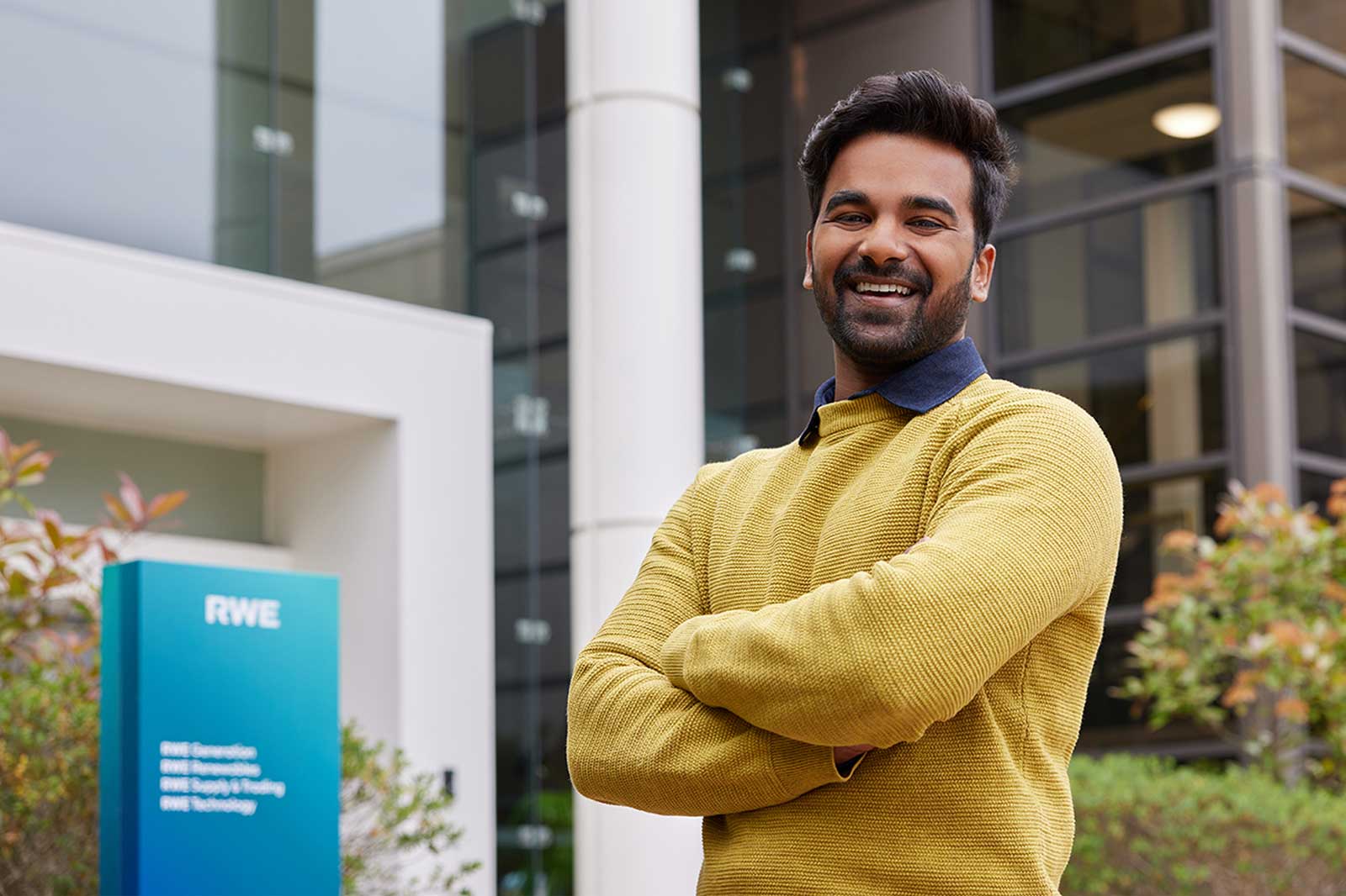 A person standing confidently with crossed arms in front of a modern building, wearing a yellow sweater and a dark collar.