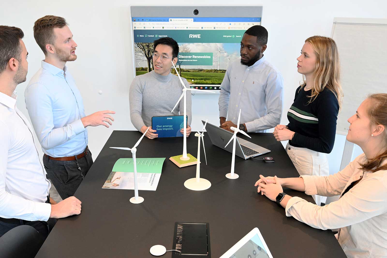 A group of professionals engaged in a discussion around a table, featuring wind turbine models and a laptop, with a presentation screen in the background.