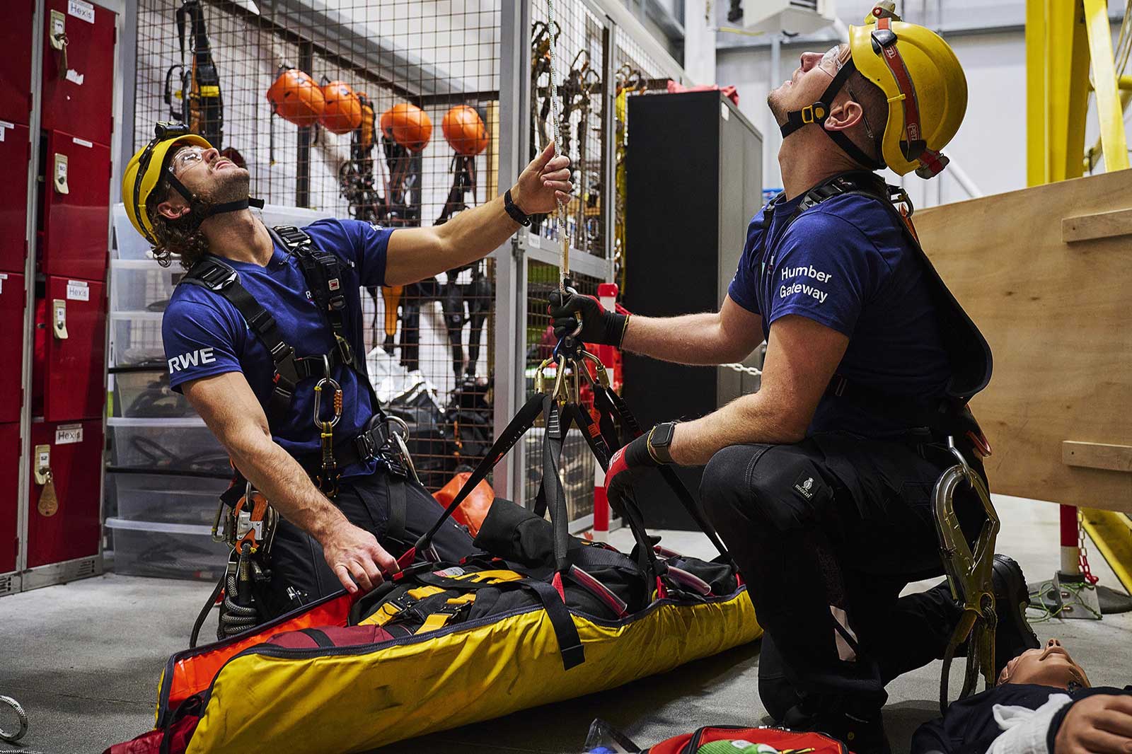 Two rescue workers in harnesses preparing equipment in a facility, with safety gear and rescue bags visible around them.