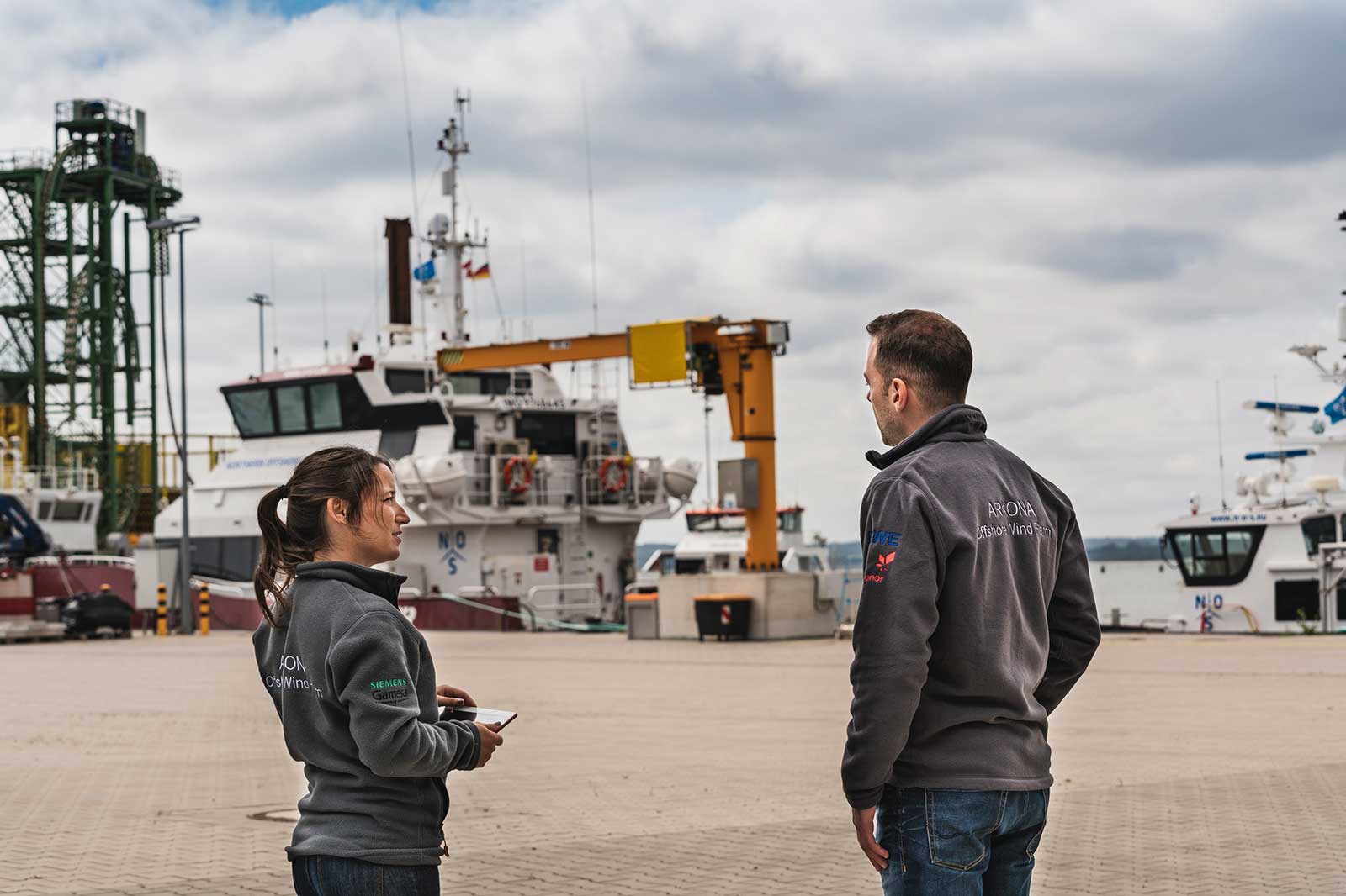 Two workers in grey jackets observe harbour activity with ships and a crane in the background, set against a cloudy sky.