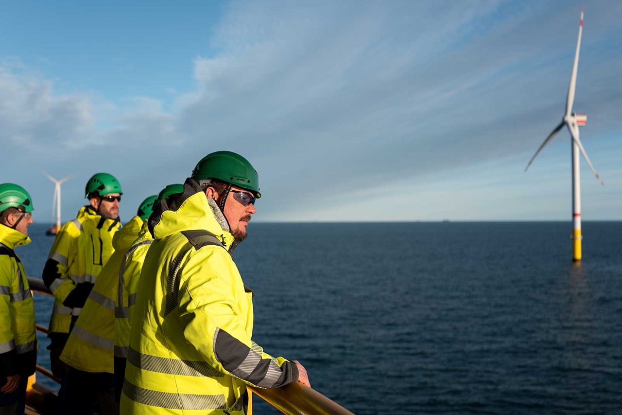 Two workers in grey jackets observe harbour activity with ships and a crane in the background, set against a cloudy sky.