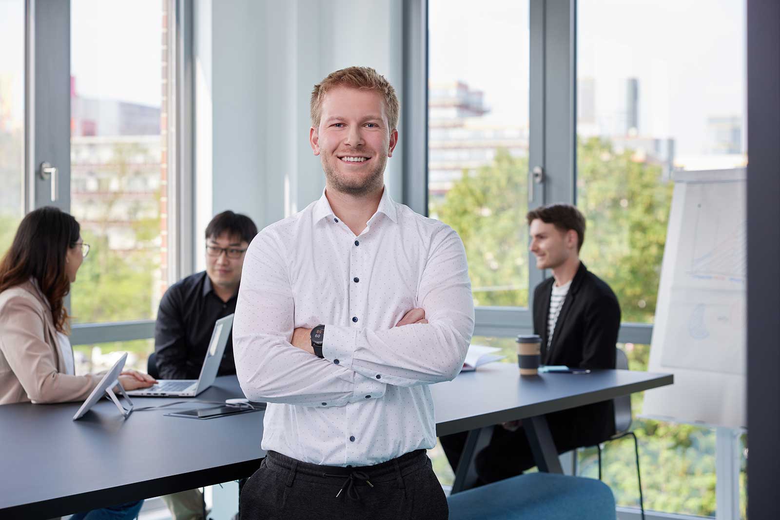 A confident man stands with arms crossed in an office, while colleagues work at a table nearby with natural light coming through windows.