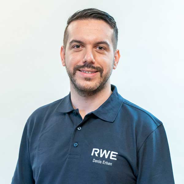 Ein Mann trägt ein dunkles Poloshirt mit RWE-Logo und steht vor einem schlichten Hintergrund.