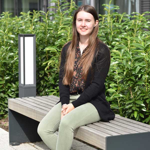 Eine Frau sitzt auf einer Holzbank, trägt einen schwarzen Cardigan und eine gemusterte Bluse, neben einer modernen Lampe, umgeben von Grün.