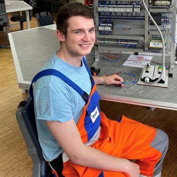 Eine Person in orangefarbener Arbeitskleidung sitzt an einem Arbeitsplatz mit sichtbaren elektrischen Komponenten und Werkzeugen auf einem Tisch.