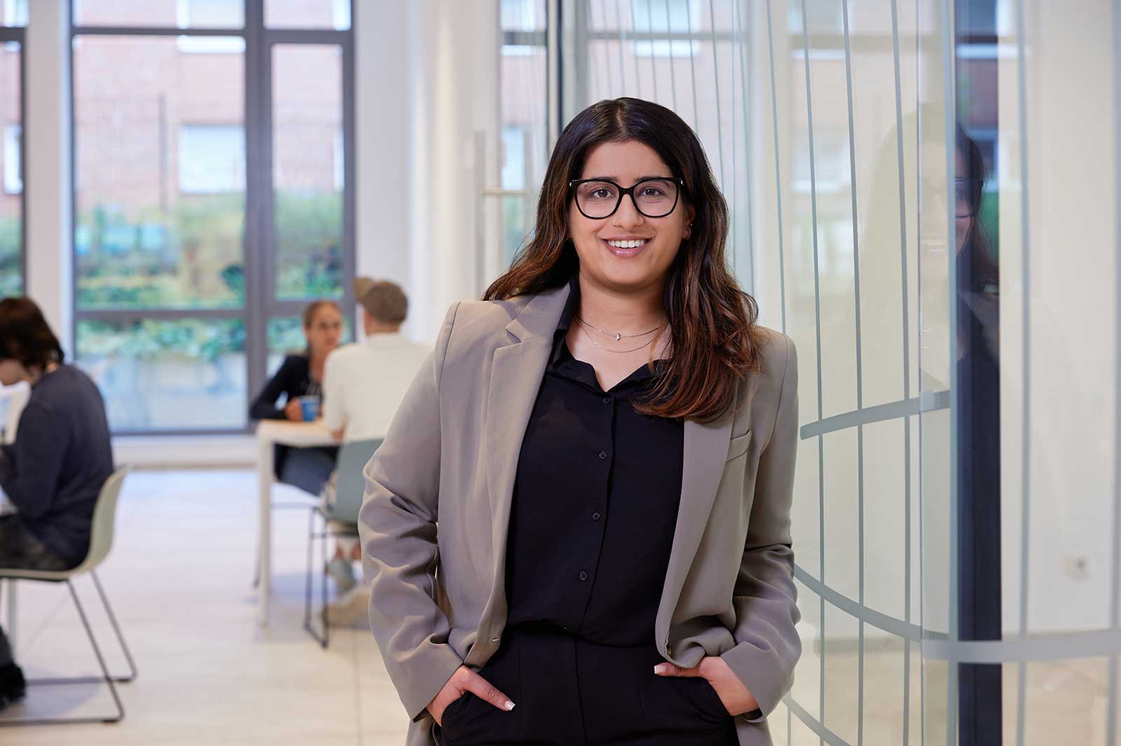 A woman in a beige blazer and black shirt stands in a modern office with large windows and a bright, open space.