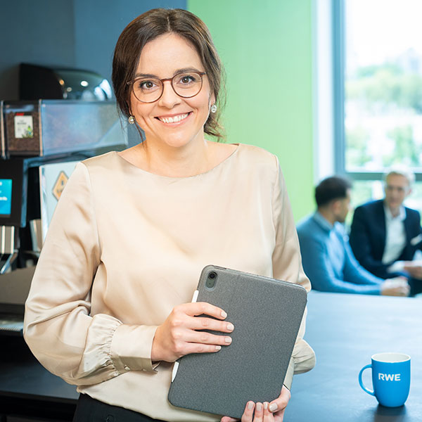 Eine Frau in einer Bluse hält ein graues Tablet, während sich eine Gruppe von Menschen im Hintergrund unterhält.