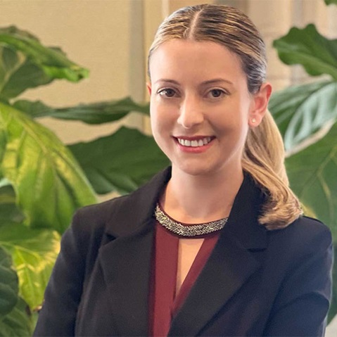 A person in a black blazer poses in front of green plants.