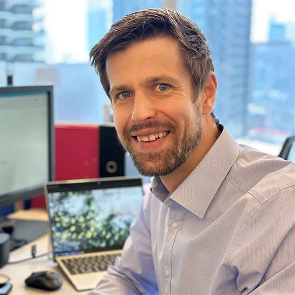 Herr in einem hellblauen Hemd sitzt an einem Schreibtisch vor Computermonitoren und einem Laptop mit einer Naturaufnahme.