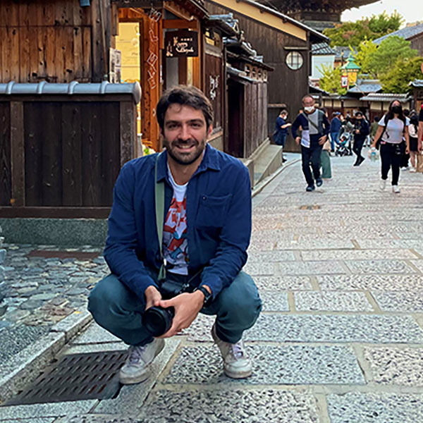 A person squatting on a cobbled street, holding a camera, surrounded by traditional wooden buildings and people walking.