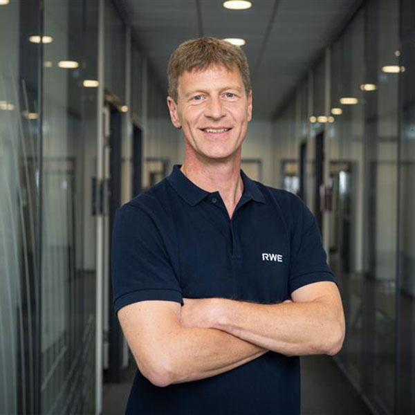 Ein Mann steht mit überkreuzten Armen in einem modernen Büroflur mit Glaswänden und hellem Licht.