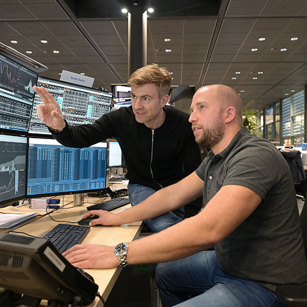 Zwei Personen analysieren Daten an einem Schreibtisch mit mehreren Bildschirmen, die Finanzinformationen zeigen.