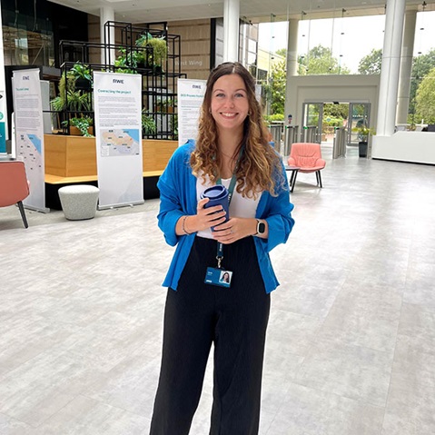 Eine Frau in einer blauen Strickjacke hält eine Kaffeetasse in einer modernen Bürolobby mit Sitzgelegenheiten und Grünpflanzen.