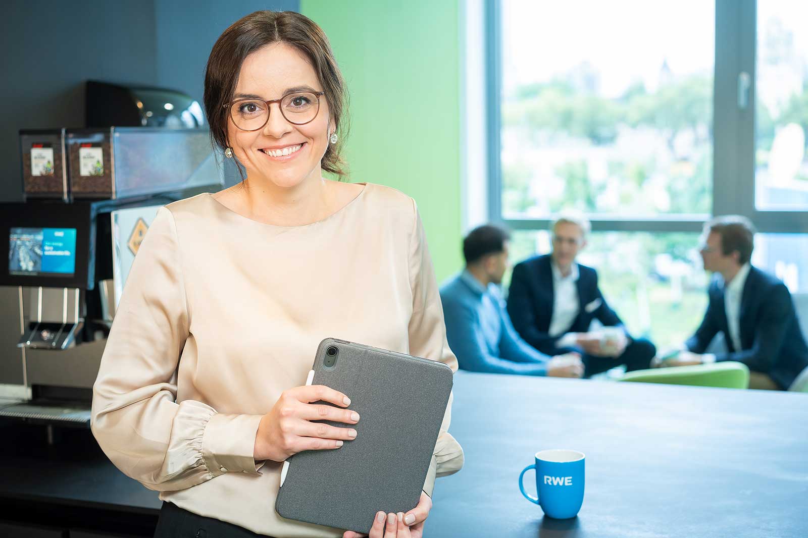 A woman holding a tablet in a modern office with a coffee mug labelled RWE in the foreground and colleagues chatting in the background.