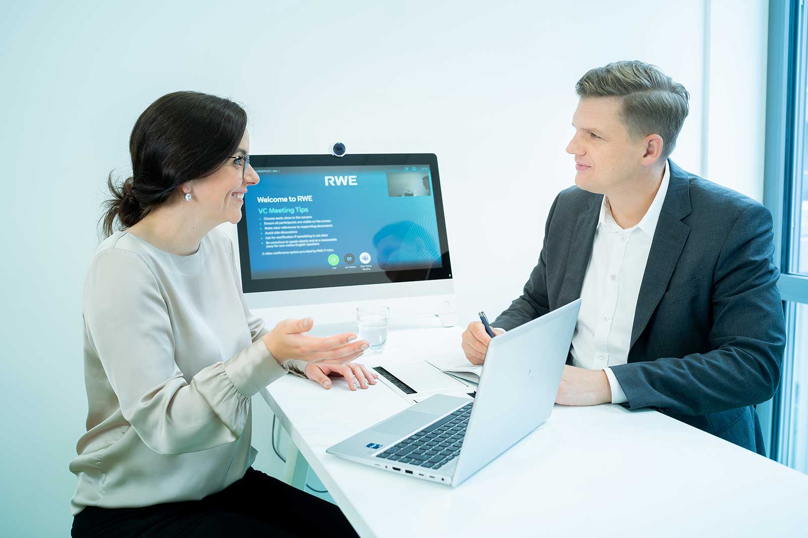 Zwei Personen sitzen an einem weißen Tisch in einem hellen Büro, eine gestikuliert, die andere schreibt an Laptops; ein Monitor zeigt „RWE“ und „Welcome to RWE“.