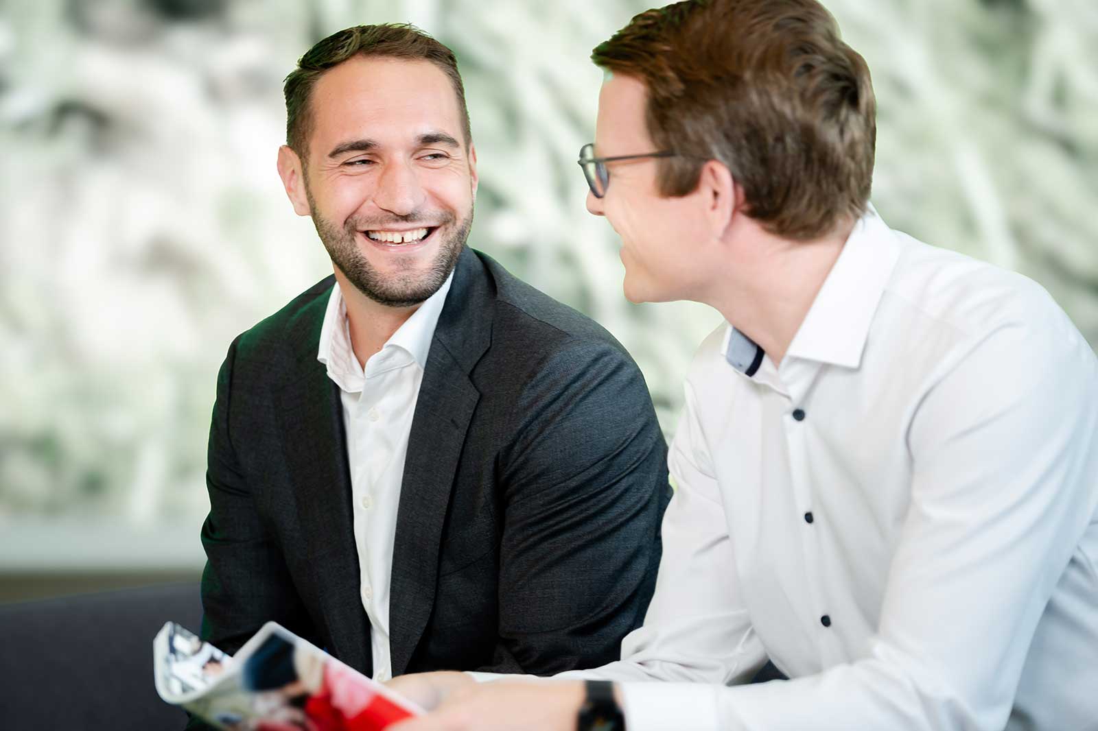 Two men in formal attire engage in conversation in a modern office with a blurred background.