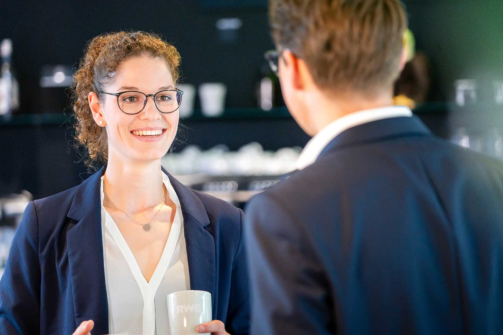 A suited individual is speaking to another person in a café with cups visible in the background.